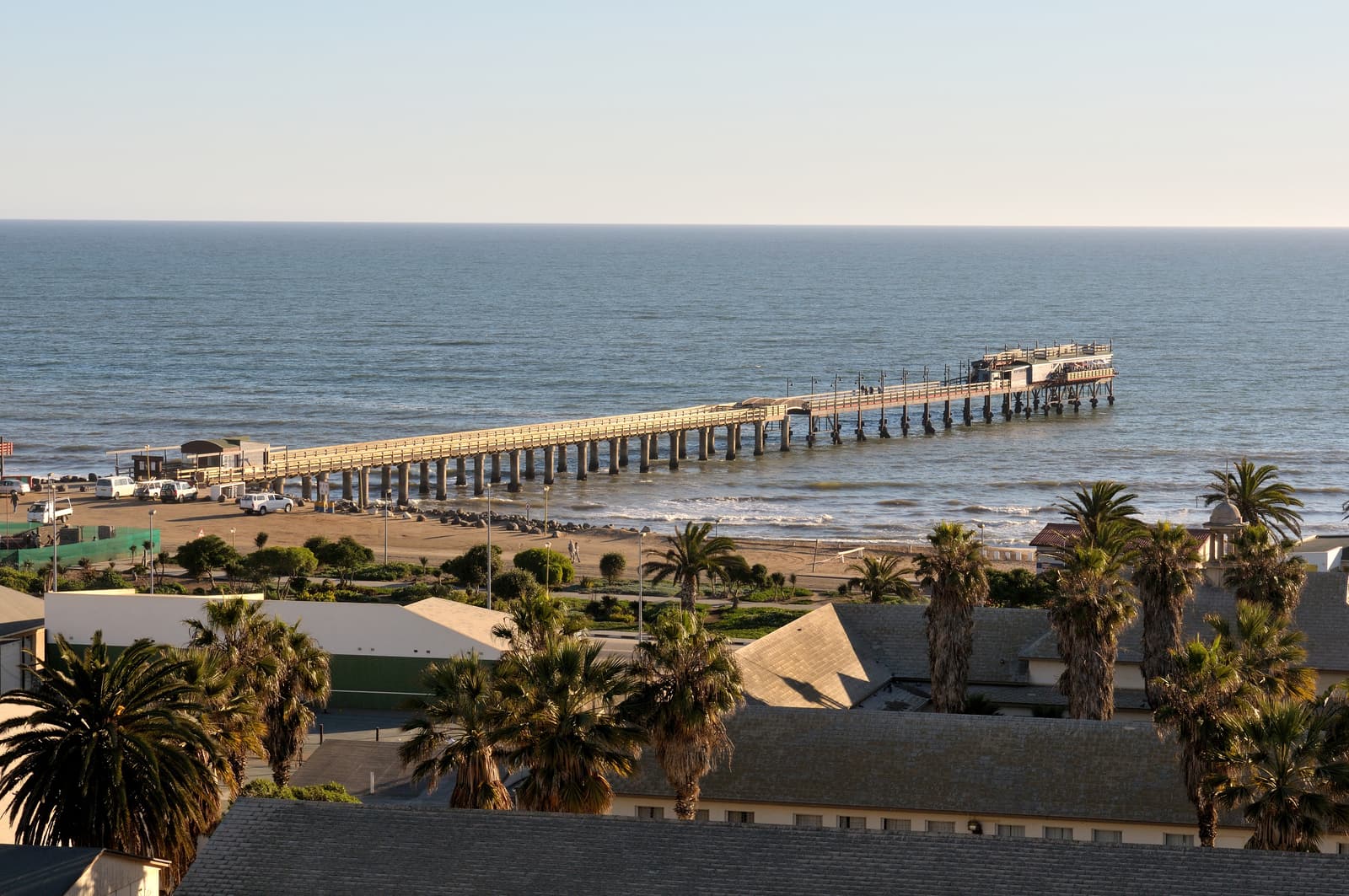Blick über Palmen und Dächer von Swakopmund auf die Jetty im Atlantik unter blauem Himmel