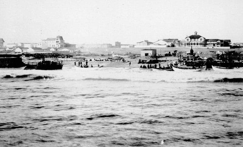 Schwarzweißfoto: offene Boote mit Menschen in der Brandung vor dem Strand von Swakopmund, im Hintergrund Wellen