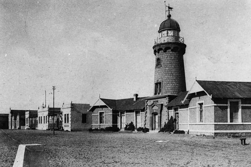 Schwarzweißfoto: schlanker Leuchtturm am Strand, daneben niedrige Gebäude, noch ohne den späteren oberen Aufbau
