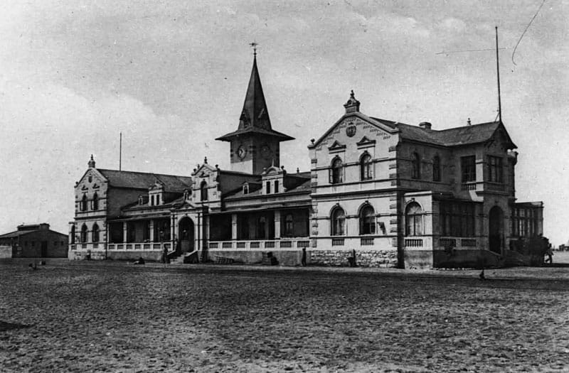 Schwarzweißfoto: langgestrecktes Bahnhofsgebäude mit Giebeln und Türmchen, davor ein freier Platz